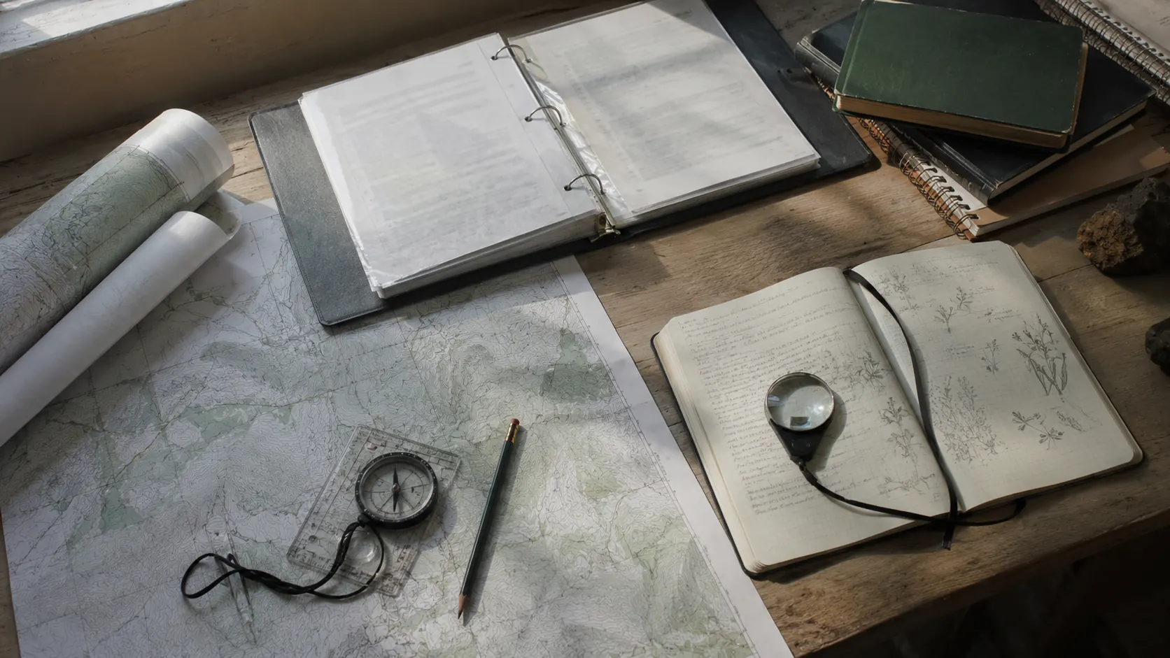 Mesa de trabajo con mapas topográficos, un expediente técnico abierto, brújula y una libreta de campo con anotaciones y dibujos de vegetación